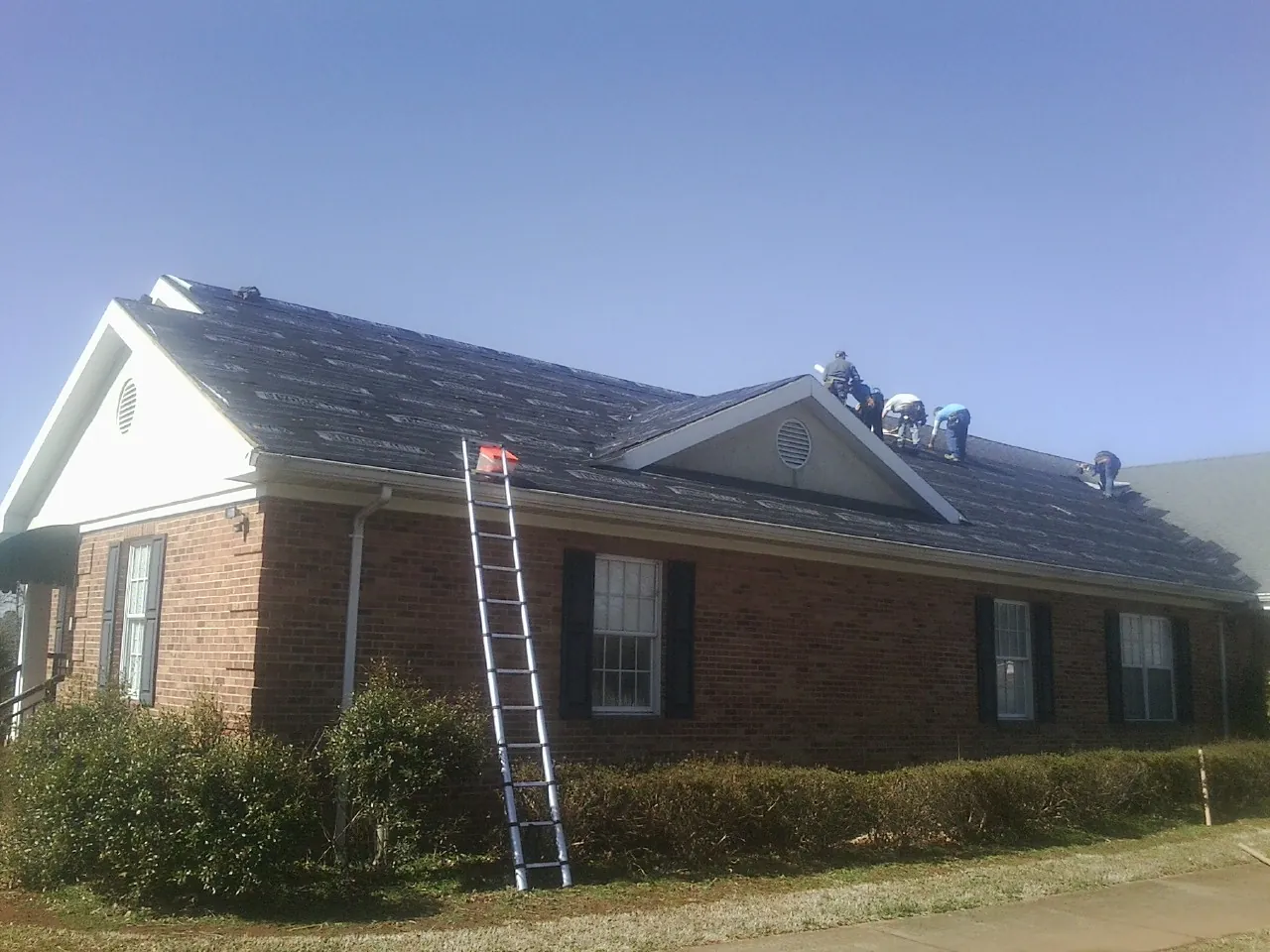 Crew tearing shingles off a brick ranch roof with a ladder set — Emergency Roof Repair work by our crew in Roan Mountain