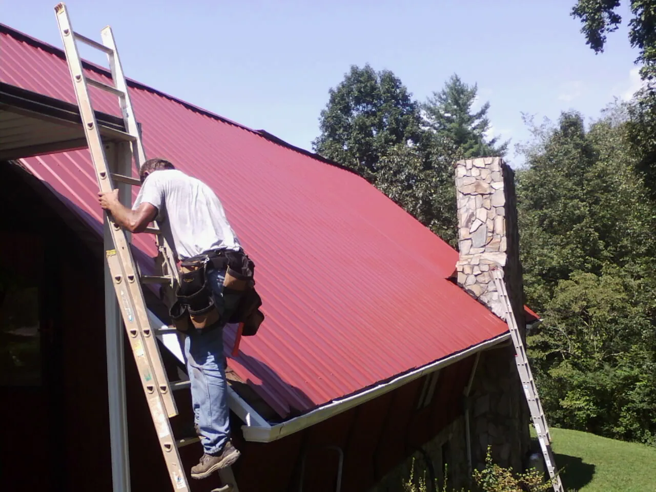 Roofer on a ladder beside a red metal roof and stone chimney — Soffit & Fascia Repair by Roofing Brothers in Roan Mountain